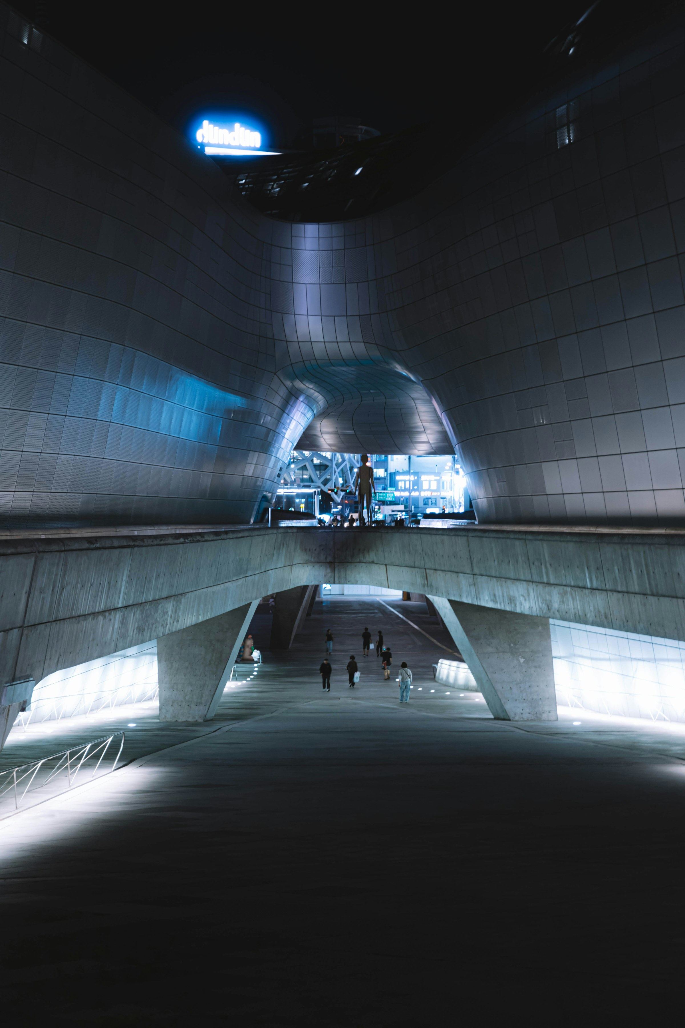A night-time tunnel beneath the curved metal-clad Dongdaemun Design Plaza in Seoul, with small figures walking toward a blue-lit exit.