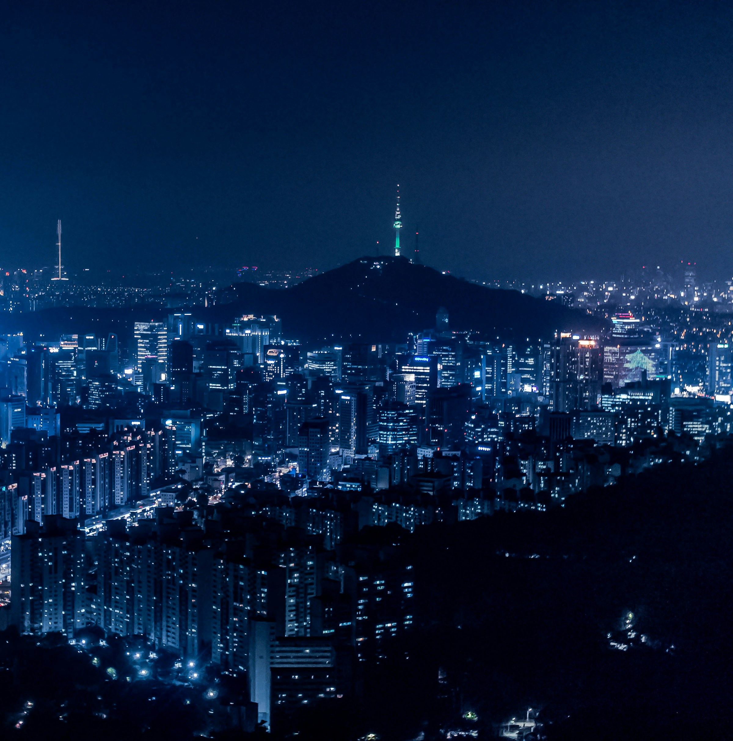 Seoul's dense night skyline spreads below Namsan hill, with N Seoul Tower lit green at its summit under a deep blue sky.