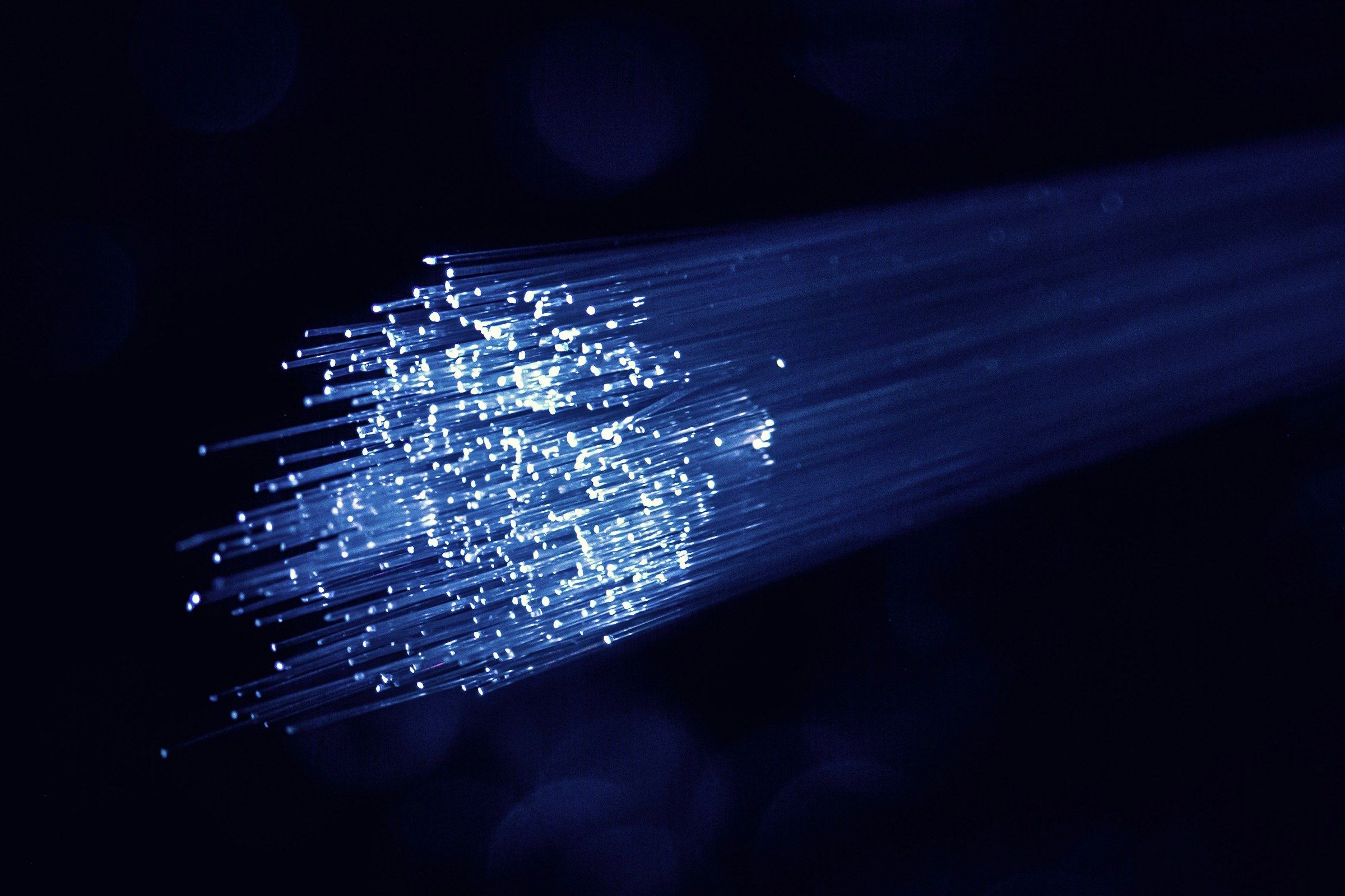 A bundle of blue-lit optical fibres fanning out from a bright tip against a dark, bokeh-soft background.