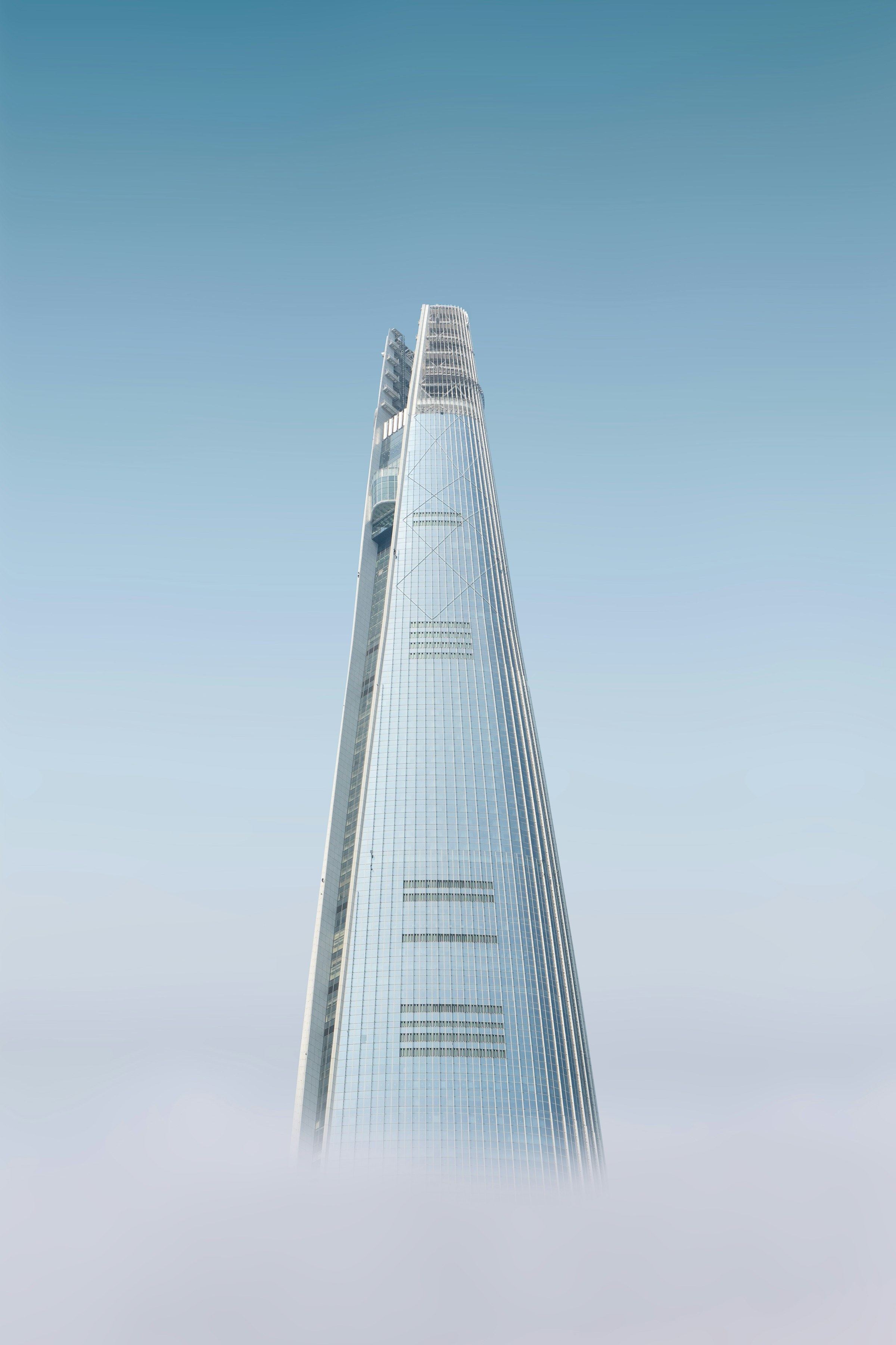 The tapered glass spire of Lotte World Tower emerges from a bank of pale cloud under a washed blue sky.